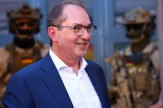 Dobrindt, leicht lächelend, mit vermuteten und bewaffneten Menschen im Hintergund