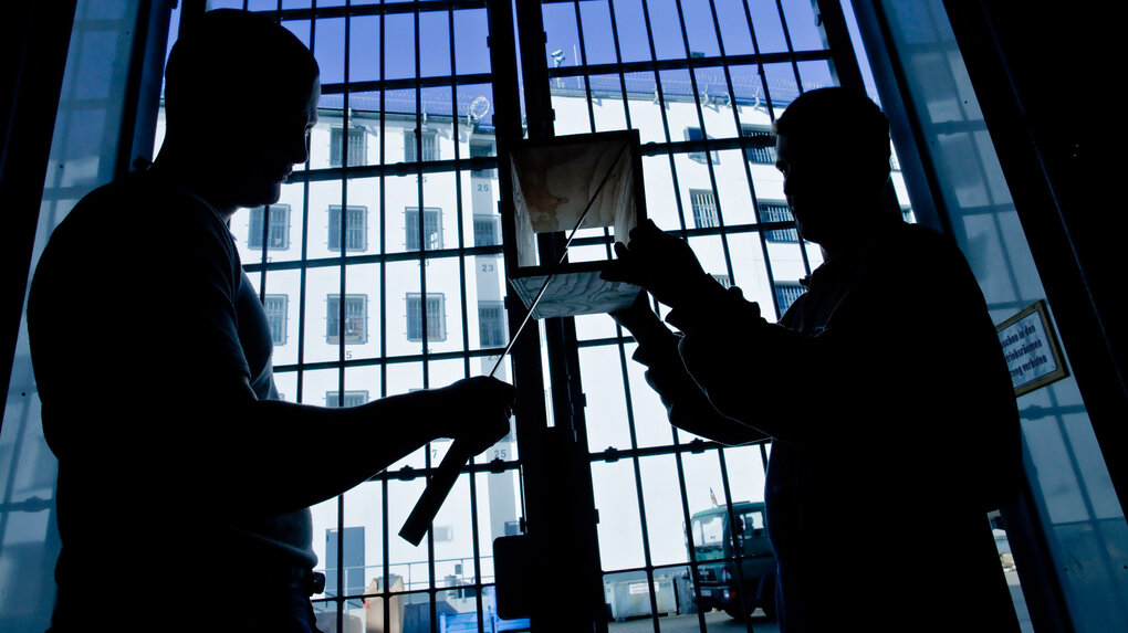 Zwei Menschen handwerken im Gegenlicht vor einem Gefängnisgitter