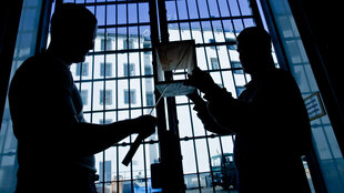 Zwei Menschen handwerken im Gegenlicht vor einem Gefängnisgitter