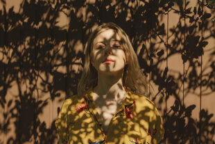 Eine Frau, die im Schatten von Bäumen steht, wobei das Sonnenlicht Schatten auf einen braunen Hintergrund wirft