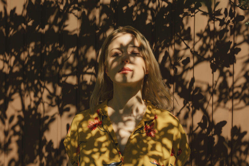 Eine Frau, die im Schatten von Bäumen steht, wobei das Sonnenlicht Schatten auf einen braunen Hintergrund wirft