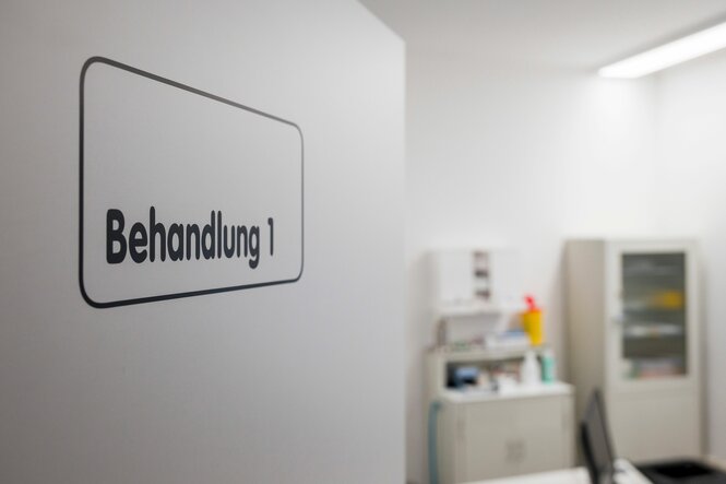 Eine offene Tür mit der Aufschrift "Behandlung 1". Im Hintergrund ein Raum mit medizinischen Utensilien
