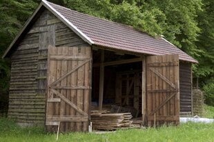 Eine gewöhnliche Holzhütte am Waldrand, eine doppelflügelige Tür ist weit geöffnet, darin liegt stapelweise Holz.