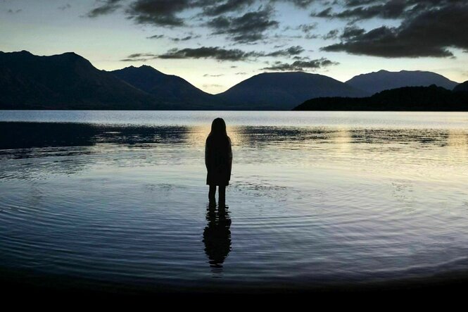 Ein junges Mädchen steht in der Abenddämmerung bis zu den Knien im See. Zu sehen isit ihre Rückenansicht