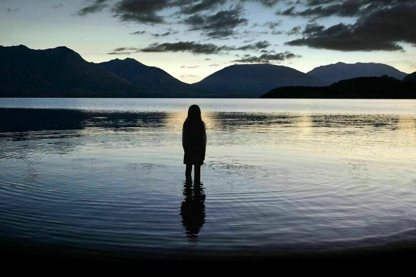 Ein junges Mädchen steht in der Abenddämmerung bis zu den Knien im See. Zu sehen isit ihre Rückenansicht