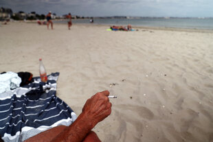 Eine Person sitzt am Strand, nur die Hand mit der brennenden Zigarette ist zu sehen