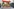 Eine Gruppe von Straßenkehrer:innen steht auf einer mobilen Konzertbühne im öffentlichen Raum. Vor der Gruppe stehen Mikrofone, die Mitglieder halten Notenblätter in der Hand, eine Person hält eine Gitarre. Am Bildrand sind Fotograf:innen und Lautsprecher zu sehen.