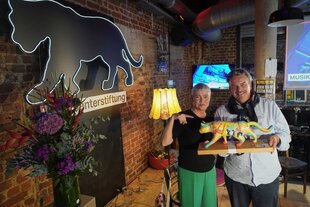 Zwei Menschen halten den Panter-Preis, eine bunte Katze auf einem Sockel, in einem Raum mit Backsteinwänden, Bühne und Bestuhlung. Auch an der Wand prangt ein Panter als Logo der taz Panter Stiftung.