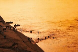 Am Ufer eines Flusses, getaucht in goldenes Abendlicht, stehen und baden zahlreiche Menschen. Boote liegen am Rand, lange Leinen sind vom Ufer ins Wasser gelegt, ein paar Menschen ziehen daran – vielleicht spielen oder fischen sie.