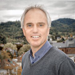 Christopher Bangert hat kurzes graues Haar, einen dunklen Pulli mit V-Ausschnitt und darunter ein Polohemd an. Er lächelt mit geöffnetem Mund und steht vor ein paar Häusern und den Ausläufern des Schwarzwalds in Freiburg.