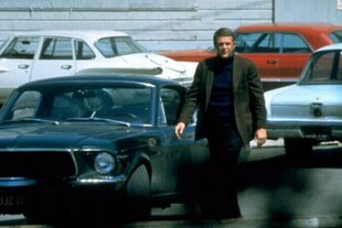 Ein Mann mit einer braunen Jacke läuft zwischen parkenden Autos in Blau-, Weiß- und Rottönen entlang.
