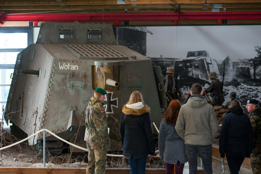 Das Bild zeigt einen alten Panzer namens "Wotan" mit einem schwarzen Kreuz und Geschützrohr, umgeben von Barrikaden und Stacheldraht. Im Hintergrund ein Foto mit Soldaten und zerstörter Landschaft. Mehrere Personen, darunter ein Soldat, betrachten den Panzer in einem Raum mit hoher Decke und Fenstern.