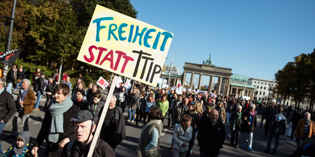 Viele Menschen auf einer Straße, einer hat ein Schild mit der Aufschrift "Freiheit statt TTIP", im Hintergrund das Brandenburger Tor (Berlin)