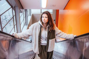 Luisa Neubauer in einer weißen Jacke auf einer Rolltreppe in einer orange-gestrichenen U-Bahn Station.