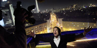 In der Nacht, ein Mann (Alexander Scheer) mit ausgebreiteten Armen auf einem Dach, hinter ihm eine Straße, dahinter ragt der Berliner Fernsehturm und das Hotel am Alexanderplatz auf