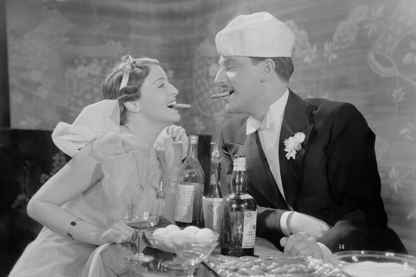Eine Frau und ein Mann in schicker Abendgarderobe stehen an einer Bar, auf der Flaschen und gekochte Eier stehen. Sie lachen und halten Knabbergebäck zwischen den Zähnen, während sie sich anschauen.