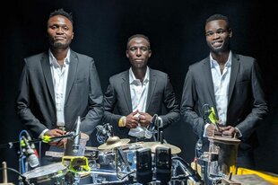 Die drei Musiker von Arsenal Mikebe stehen vor einem vielfältigen Schlagwerkset, über dem Mikrofone angebracht sind.