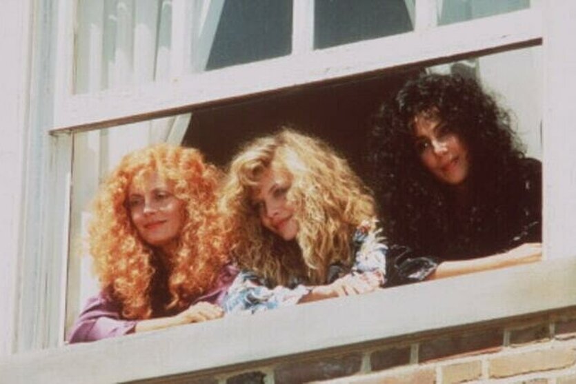 Die drei Hexenfiguren aus dem Film "The Witches of Eastwick", gespielt von Susan Sarandon, Michelle Pfeiffer und Cher, schauen aus dem Fenster eines Hauses nach unten und lachen.