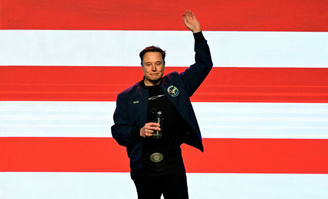 Elon Musk steht vor einer Wand mit roten und weißen Streifen. Er hebt den linken Arm in die Luft.