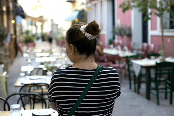 Setareh E. steht in einer kleinen Gasse mit Außengastronomie. Sie trägt ein Ringel T-Shirt, hat die Haare hochgesteckt und ist nur von hinten zu sehen
