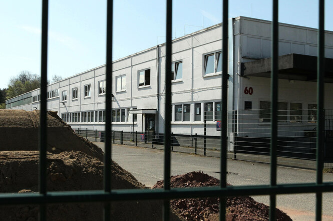 Blick durch ein Gitter auf ein langgestrecktes Gebäude das aus Containern besteht und mit einem Drahtzaun umgeben ist