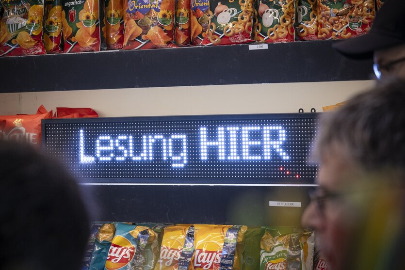 Ein LED-Bildschirm mit den Worten "Lesung hier" steht zwischen zwei Regalen mit Chipstüten.