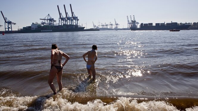 Zwei menschen baden in der Elbe, im Hintergrund sind Containerterminals des Hamburger Hafens zu sehen