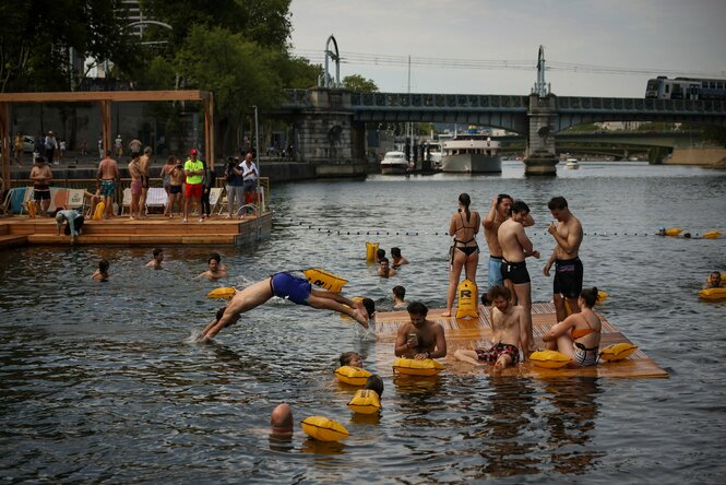 Viele Menschen stehen von einem Ponton, manche schwimmen in der Seine, man sieht städtisches Ambiente