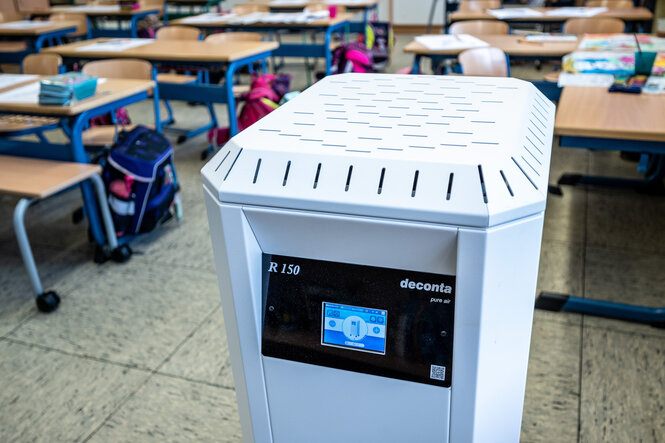 Zu sehen ist ein Klassenzimmer mit Luftfilter im Vordergrund und auf den Unterricht bereiten Tischen