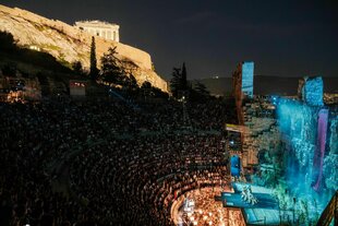 Die Freilichtbühne des Odeon Theaters am Fuße der Akropolis mit vielen Zuschauern und einer blau leuchtenden Bühne am Abend