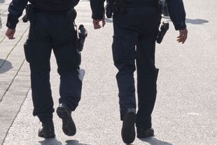Der Bildausschnitt zeigt die Bein- und Bauchpartie zweier Polizisten in Vollmontur und mit Waffengurt, die mit dem Rücken zur Kamera gewandt einen Bürgersteig entlanggehen. Die Sonne scheint.