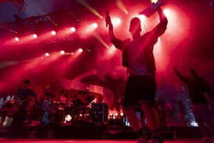 Ein Mensch steht im roten Rampenlicht einer Bühne mit gehobenen Händen und Mikrofon, dahinter eine Band mit Bassgitarre, Schlagzeug und Trompete.