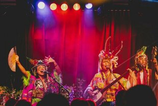 Ein sehr farbenfrohes Bühnenbild einer Musikgruppe namens Mitsune während eines Konzertes. Drei Musiker:innen sind zu sehen, alle tragen aufwendige, leuchtende Kostüme mit markanten Kopfbedeckungen. Eine Musikerin in der Mitte hält einen Fächer, während sie singt. Eine weitere Musikerin spielt ein Zupfinstrument (ähnlich einer Shamisen), und ganz rechts ist ein Musiker mit einem Kontrabass zu sehen. Die Bühne ist in leuchtendes Rot und Blau getaucht, im Vordergrund sind die Silhouetten des Publikums erkennbar.