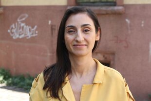Ein Porträtfoto zeigt Anahita Azizi, Protagonistin des Kontext-Artikels „Es ist so unheimlich still“. Die jüngere Frau steht in einer ortslosen Stadtlandschaft, trägt ein gelbes Hemd und schulerzlage schwarze Haare. Sie guckt entschlossen, fast schon mit einem leichten Lächeln in die Kamera.