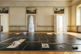 Ein eleganter Raum mit weißem Kamin, Gemälden an der Wand und einem Boden mit schwarzen Fliesenplatten, auf denen Kunstobjekte wie Schuhe und Bücher platziert sind.