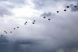 Eine lange Reihe von Fallschirmspringern fliegt vor einem grau verhangenen Himmel nach unten
