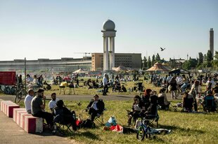 Tempelhofer Feld, viele Menschen sitzen im Gras, grillen, quatschen, sitzen auf Campingstühlen, im Hintergrund das Flughafengebäude
