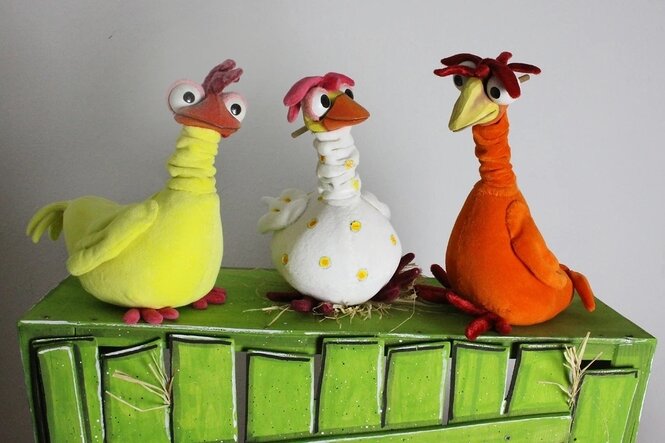 Drei bunte Stoffhühner (gelb, weiß, orange) sitzen auf einem grünen Kasten