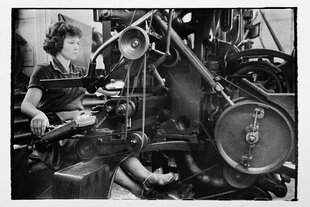 Schwarz-weiss Foto einer jungen Frau, die an einer Druckmaschine sitzt, sie schaut konzentriert