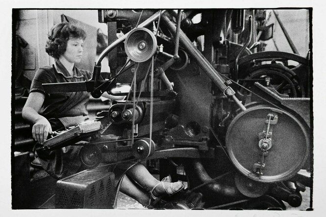 Schwarz-weiss Foto einer jungen Frau, die an einer Druckmaschine sitzt, sie schaut konzentriert