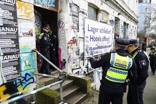 Ein Polizist in Uniform steht vor einem urbanen Gebäude mit Graffiti, Treppen und Schildern, während eine Menschenmenge bei einer Demonstration mit Postern und Solidaritätsbotschaften versammelt ist.