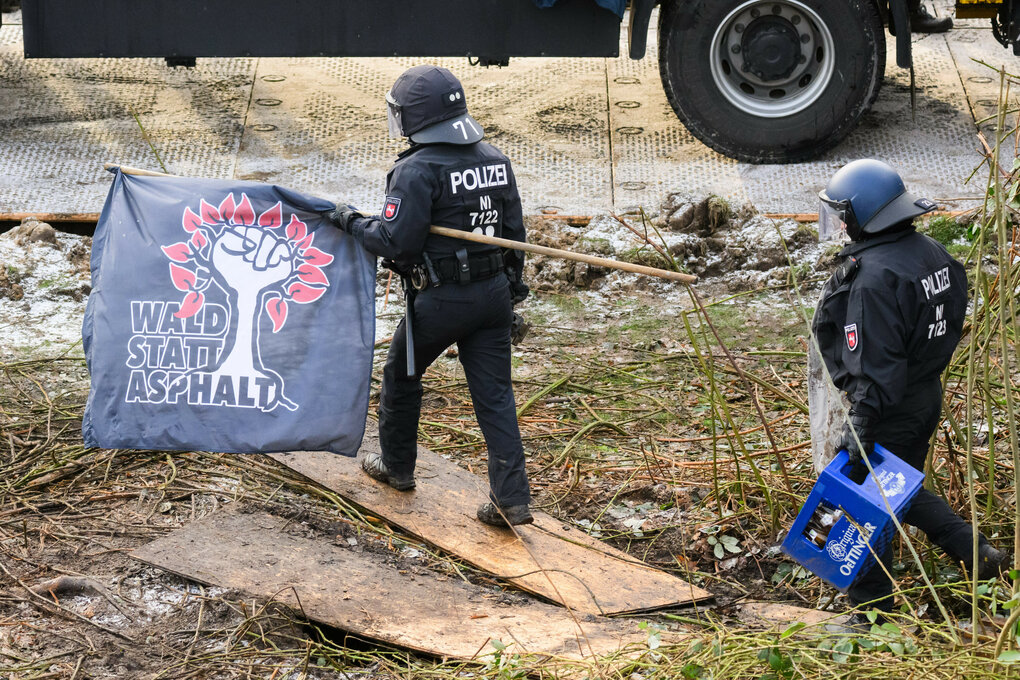 Zwei Polizisten in Uniform tragen ein Banner mit der Aufschrift "WALD STATT ASPHALT" und einem Baum-Symbol, während sie über Holzplanken gehen. Ein Polizist hält eine Kiste Bier.