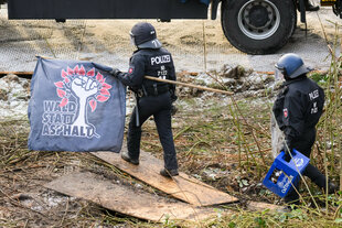 Zwei Polizisten in Uniform tragen ein Banner mit der Aufschrift "WALD STATT ASPHALT" und einem Baum-Symbol, während sie über Holzplanken gehen. Ein Polizist hält eine Kiste Bier.