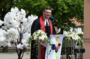 Pfarrer Alexander Stojanowič hält eine Rede. An seinem Pult ist eine Trans Pride Flagge angebracht