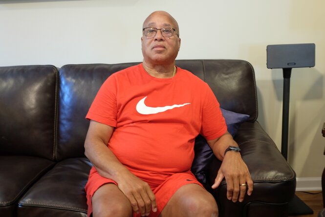 Harold Delancey sitzt in orangefarbener Sportkleidung auf einem Ledersofa