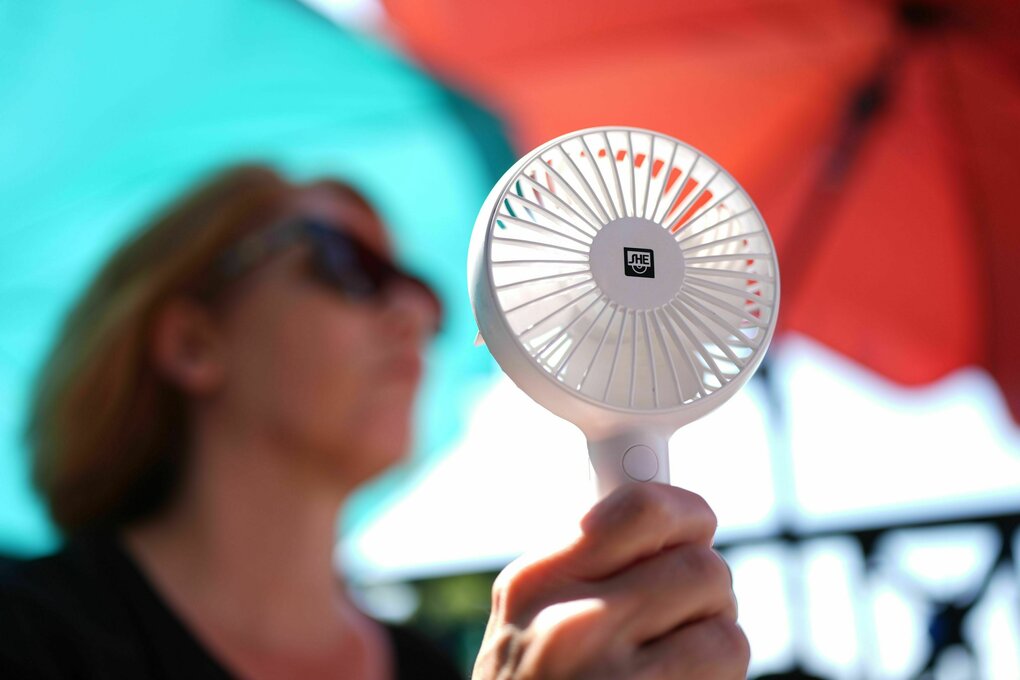 Eine Frau kühlt sich mit einem Handventilator unter Sonnenschirmen auf einem Balkon in der Mittagssonne ab