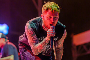 Der Sänger Frank Carter singt beim Sex-Pistols-Konzert in Berlin. Er ist verziert mit zahlreichen Tätowierungen, darunter auch an den Armen und am Hals, singt leidenschaftlich in ein Mikrofon. Er hat kurz geschnittenes, hellgrün gefärbtes Haar und Piercings im Gesicht. Im Hintergrund ist undeutlich ein weiterer Musiker zu sehen.
