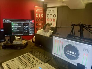 Ein Mann sitzt an einem Schreibtisch und hinter einem Mikro. Vor ihm ein anderer Tisch mit vielen Reglern vom Radio und zwei großen Bildschirmen. Hinter dem Mann eine rote Wand und eine graue Wand. Es ist ein Studio des Radiosenders.