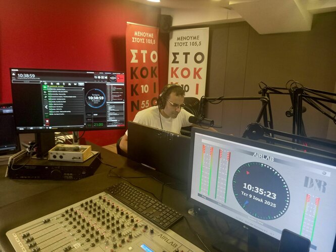 Ein Mann sitzt an einem Schreibtisch und hinter einem Mikro. Vor ihm ein anderer Tisch mit vielen Reglern vom Radio und zwei großen Bildschirmen. Hinter dem Mann eine rote Wand und eine graue Wand. Es ist ein Studio des Radiosenders.