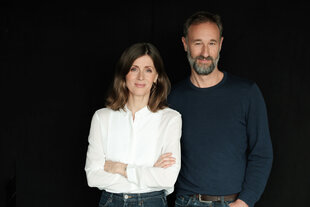 Ein Porträtfoto von der taz-Chefs Aline Lüllmann und Andreas Marggraf. Aline Lüllmann, links im Bild, trägt eine weiße Bluse und Jeans, ihre Arme sind verschränkt. Andreas Marggraf, rechts im Bild, hat einen Bart und trägt ein dunkles Langarmshirt und Jeans. Beide blicken direkt in die Kamera vor einem dunklen Hintergrund.